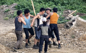 six workers carrying a person between them on a rocky construction site with dirt and rubble